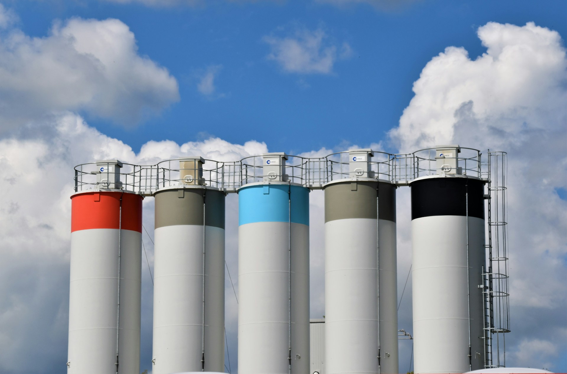 Cinque di quelle che sembrano taniche di stoccaggio industriali sono colorate verso la cima di grigio, azzurro e rosso, creando un piacevole effetto visivo