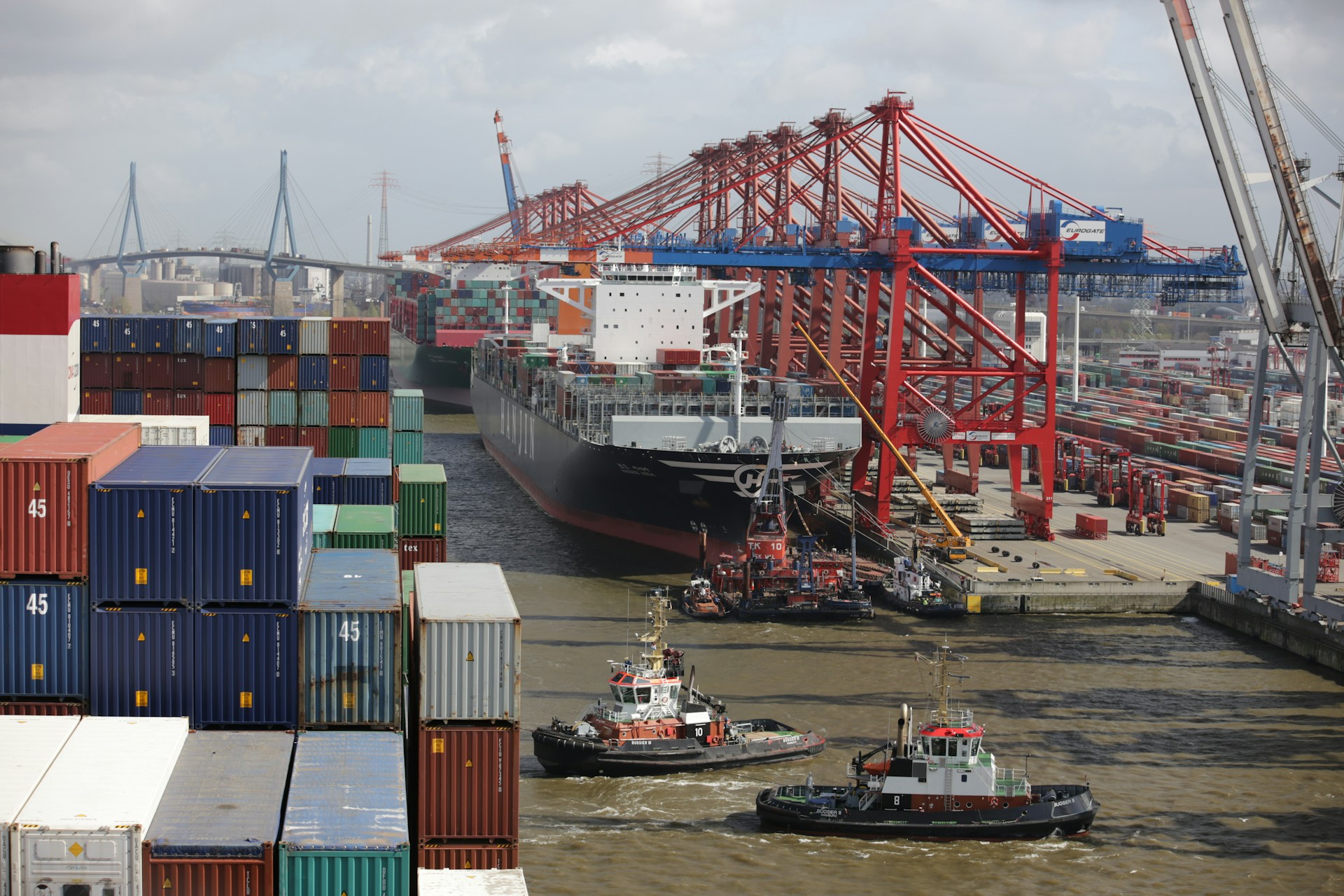 Due rimorchiatori operano nelle acque di un porto industriale. Sullo sfondo, mercantili, gru industriali e container. Si tratta proprio di supply chain.